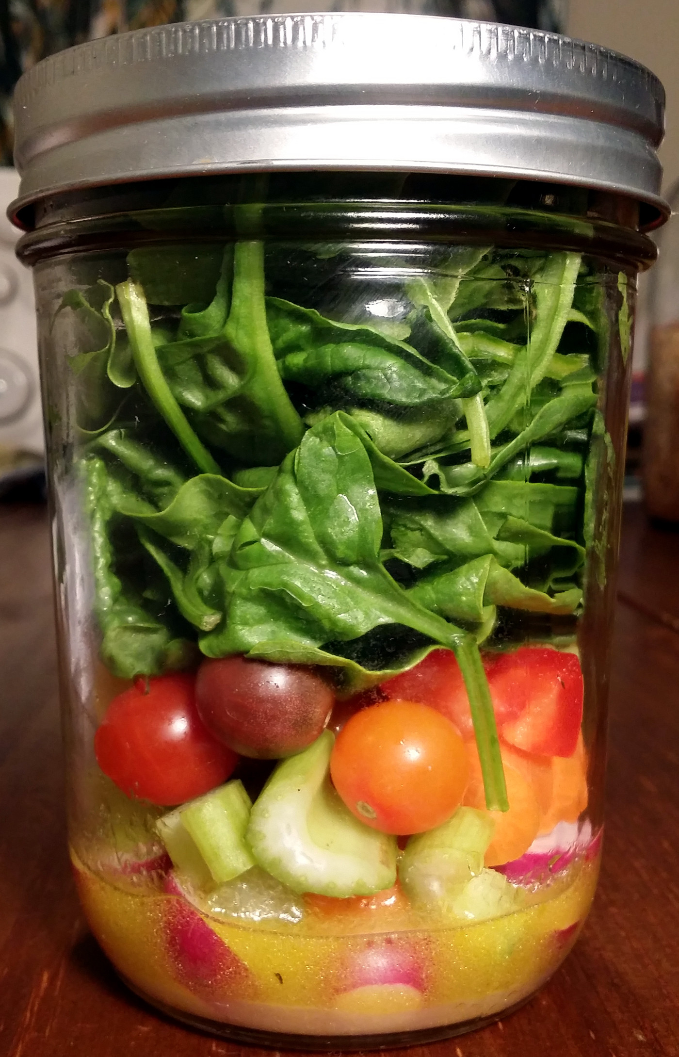 Vegetables and spinach are packed in a jar with salad dressing in the bottom. The jar is sitting on a wooden table.