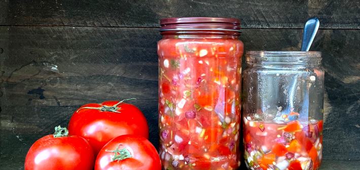 Three tomatoes and two jars of salsa on a brown wooden background. One larger jar is full. The other smaller jar is half full. The lid is off and a spoon is sitting in the jar.