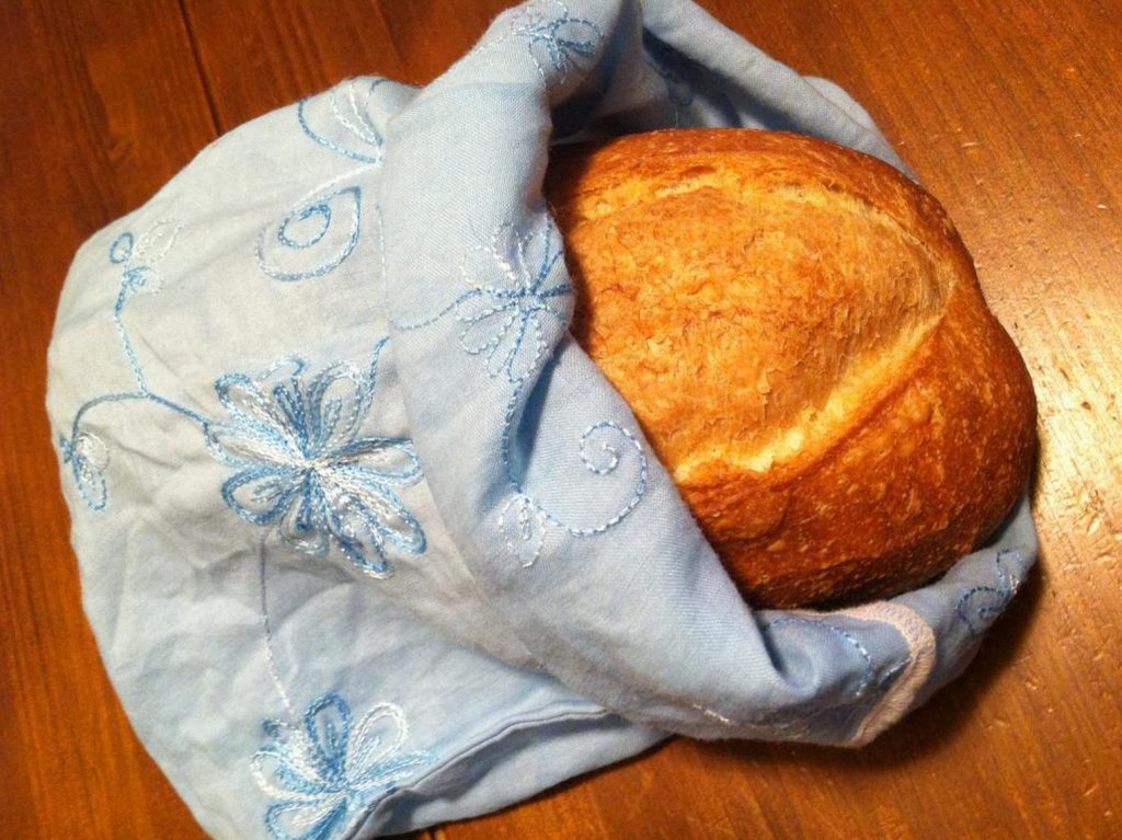 A whole loaf of fresh white bread inside a light blue cloth produce bag. The loaf is sitting on a brown wooden background.