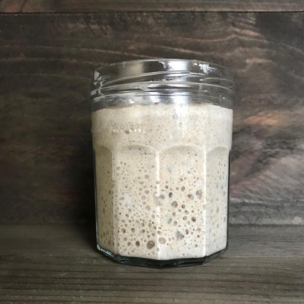 A jam jar of sourdough starter filled with bubbles. The jar is sitting on a dark brown wooden background.
