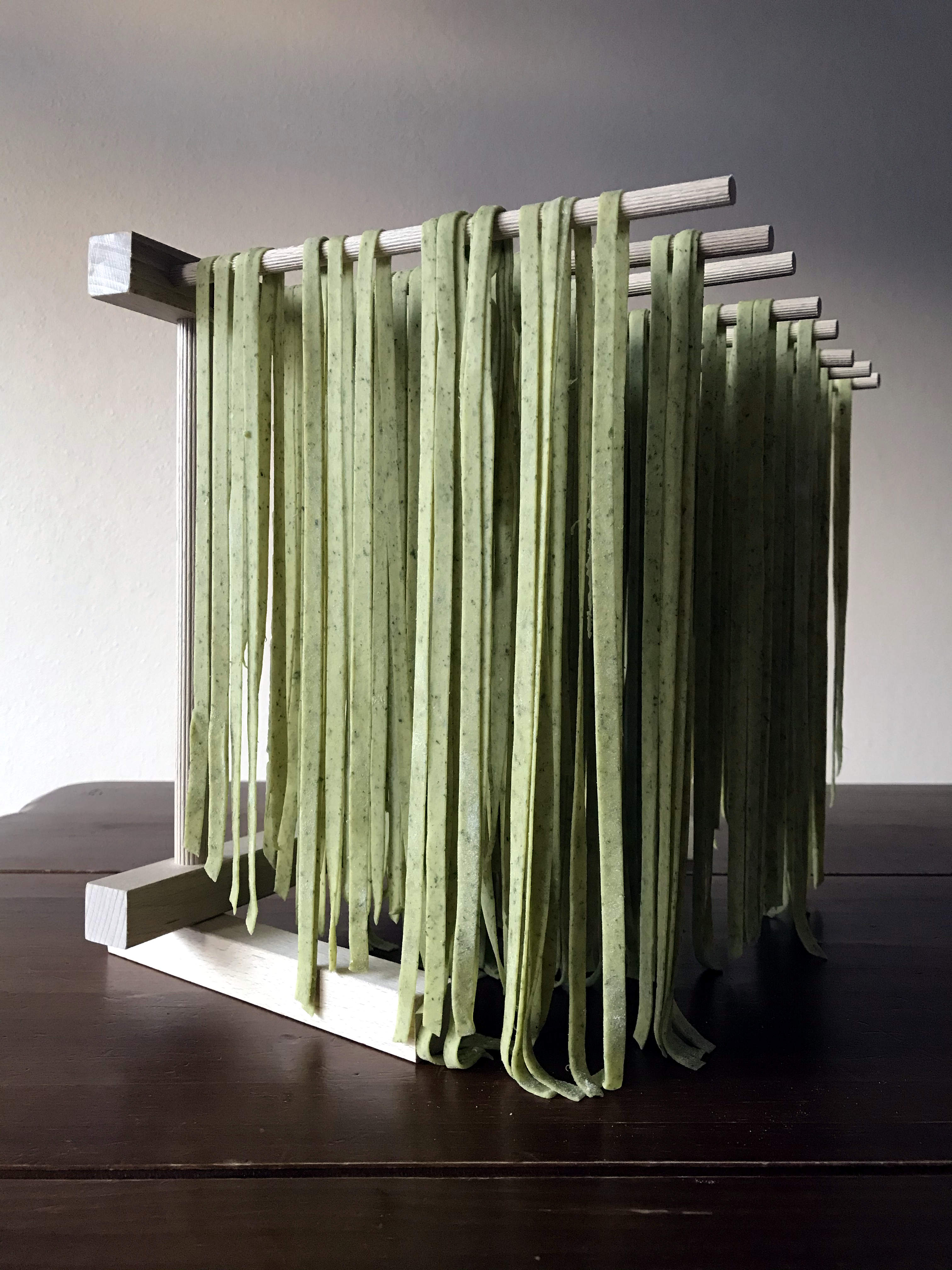 Green pasta noodles hang on a drying rack. The rack is sitting on a dark wooden table.