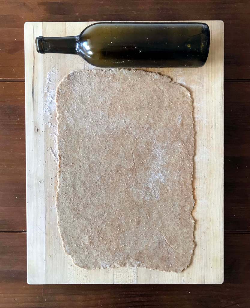 Dough rolled out on a pale wooden cutting board. A dark green wine bottle is sitting above the dough to demonstrate that it can be used as a rolling pin. The wooden board sits on a dark wooden background.