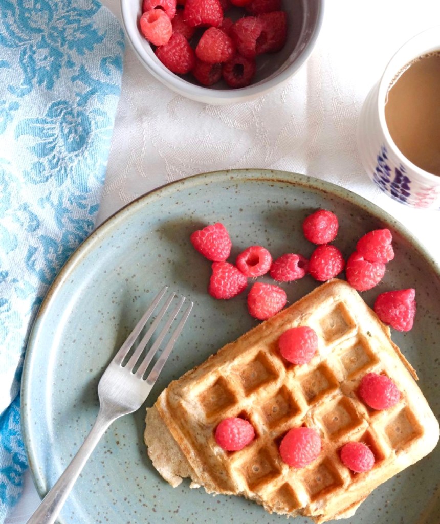 A plate has a waffle on it topped with ripe fresh raspberries. A bowl of raspberries and cup of tea sits nearby.