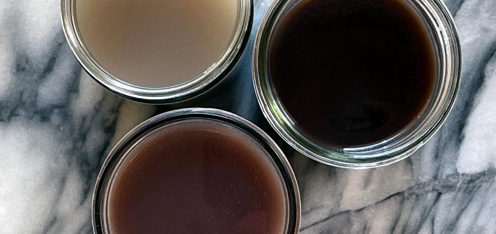 Three glass jars on a marble background, containing bean broth. They are pinto bean broth, black bean broth and kidney bean broth.