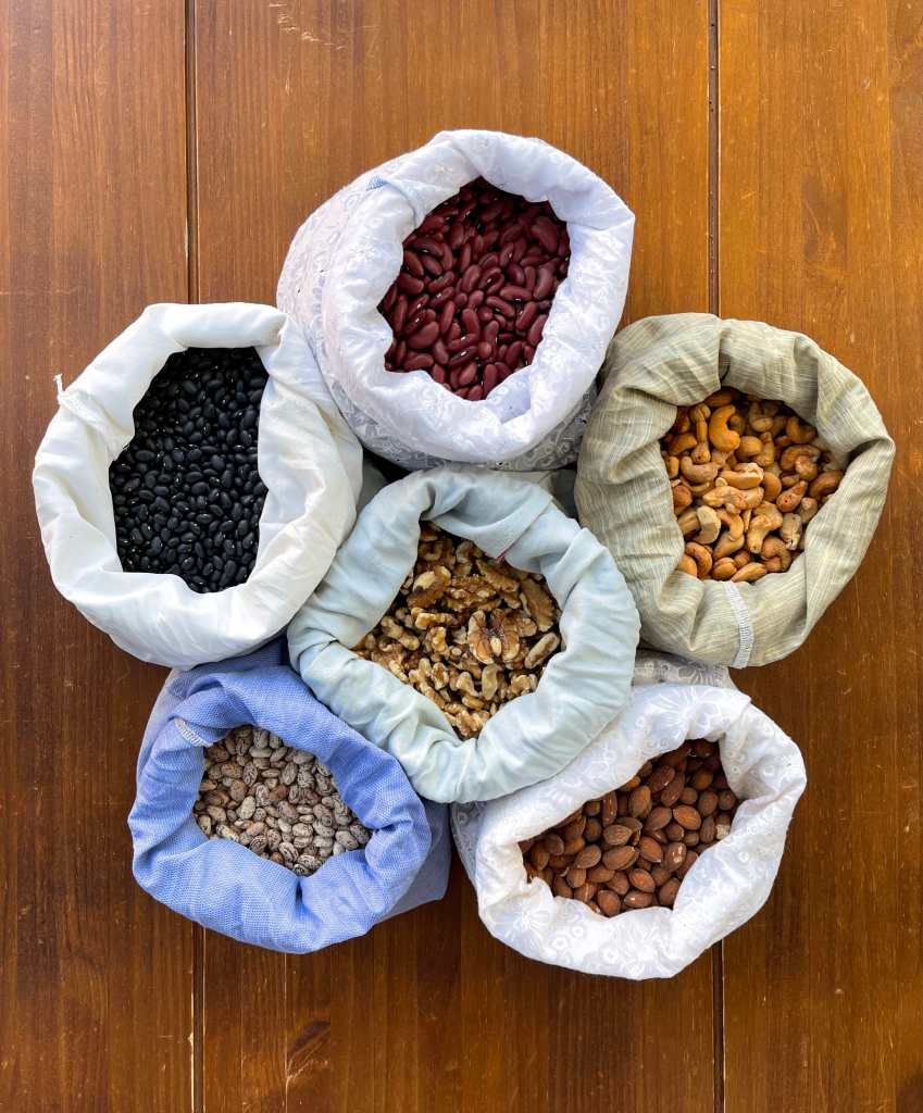 6 open cloth produce bags arranged on a wooden tabletop. The bags contain black beans, kidney beans, cashews, walnuts, pinto beans and almonds. Beans are a delicious component in frugal dinners.