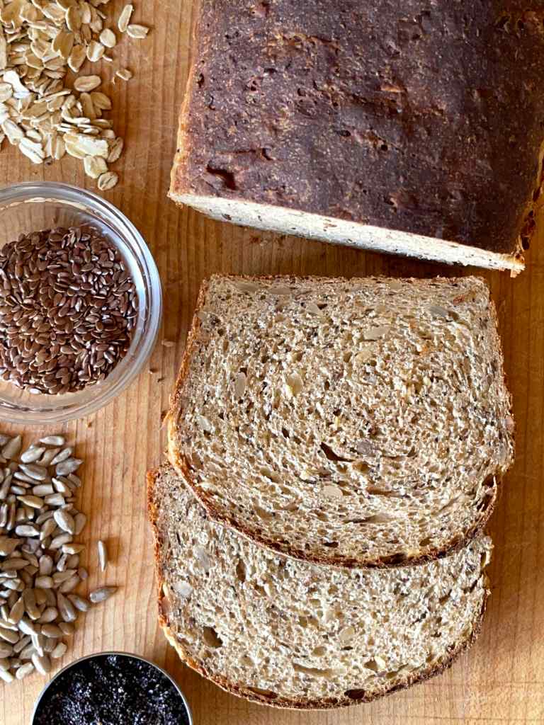 multigrain sourdough discard bread sliced on a wooden cutting board, surrounded by the various seeds that go into it