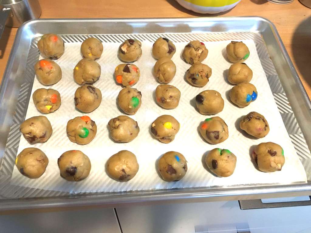 Balls of raw cookie dough before baking, resting on parchment paper on an aluminum cookie sheet. They dough balls are filled with chopped up Halloween candy.