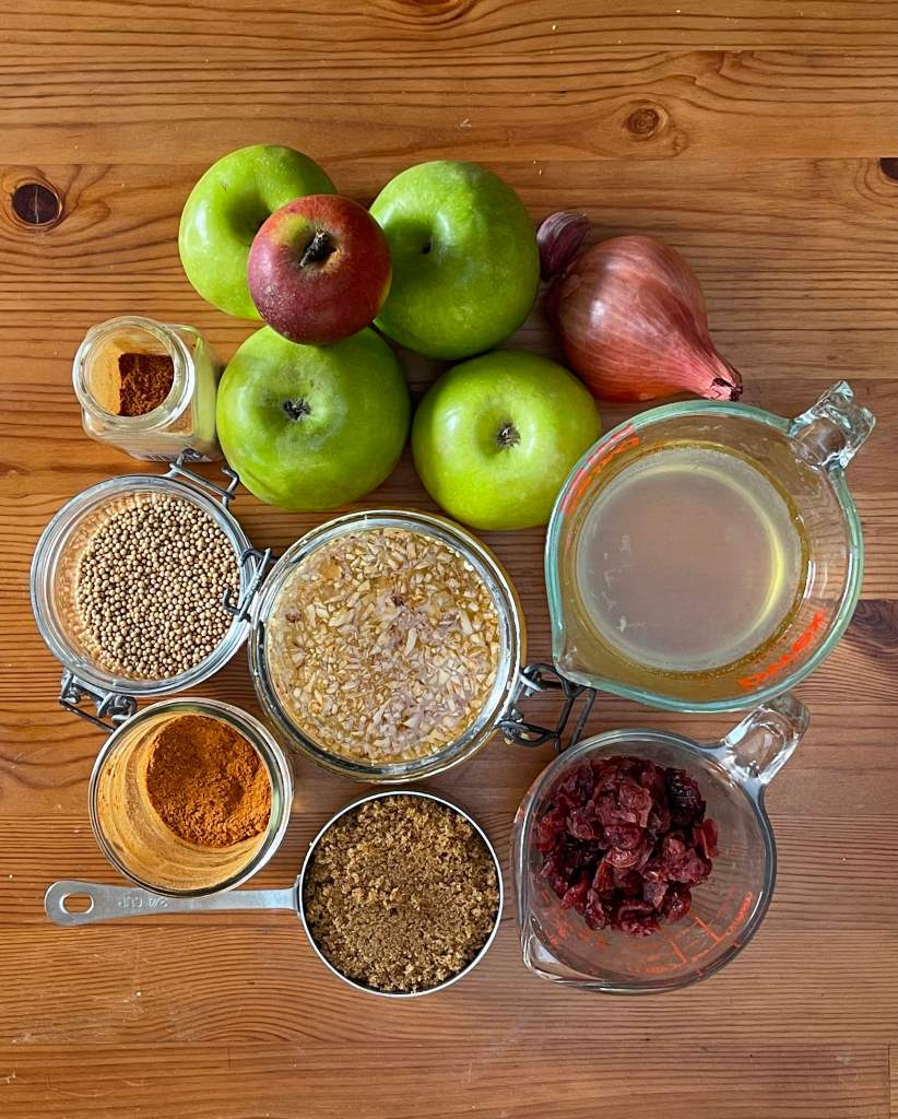 Apples, shallot, garlic, dried spices, minced ginger in a jar, vinegar, brown sugar and cranberries, all on a wooden background. These ingredients will go into chutney. 