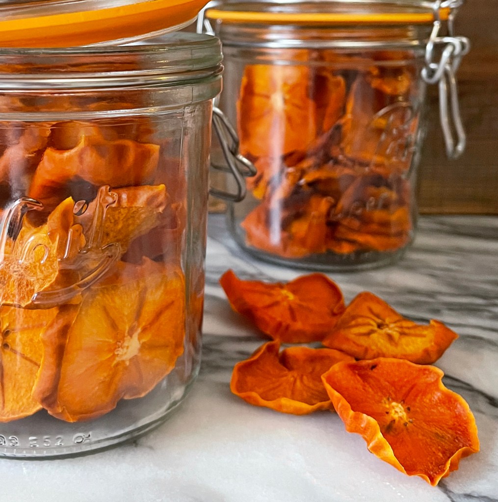Two gifts in jars of dried persimmon slices. The jars and four dried slices of persimmons are sitting on a gray and white marble top.