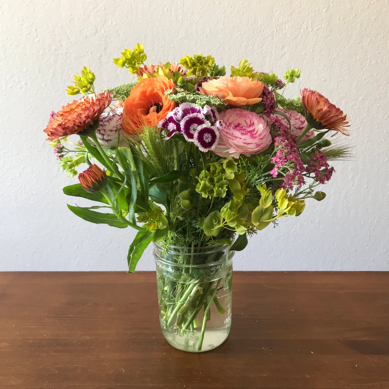 Bouquet of mixed flowers in a glass jar for a vase. The vase is sitting on a dark wooden table.