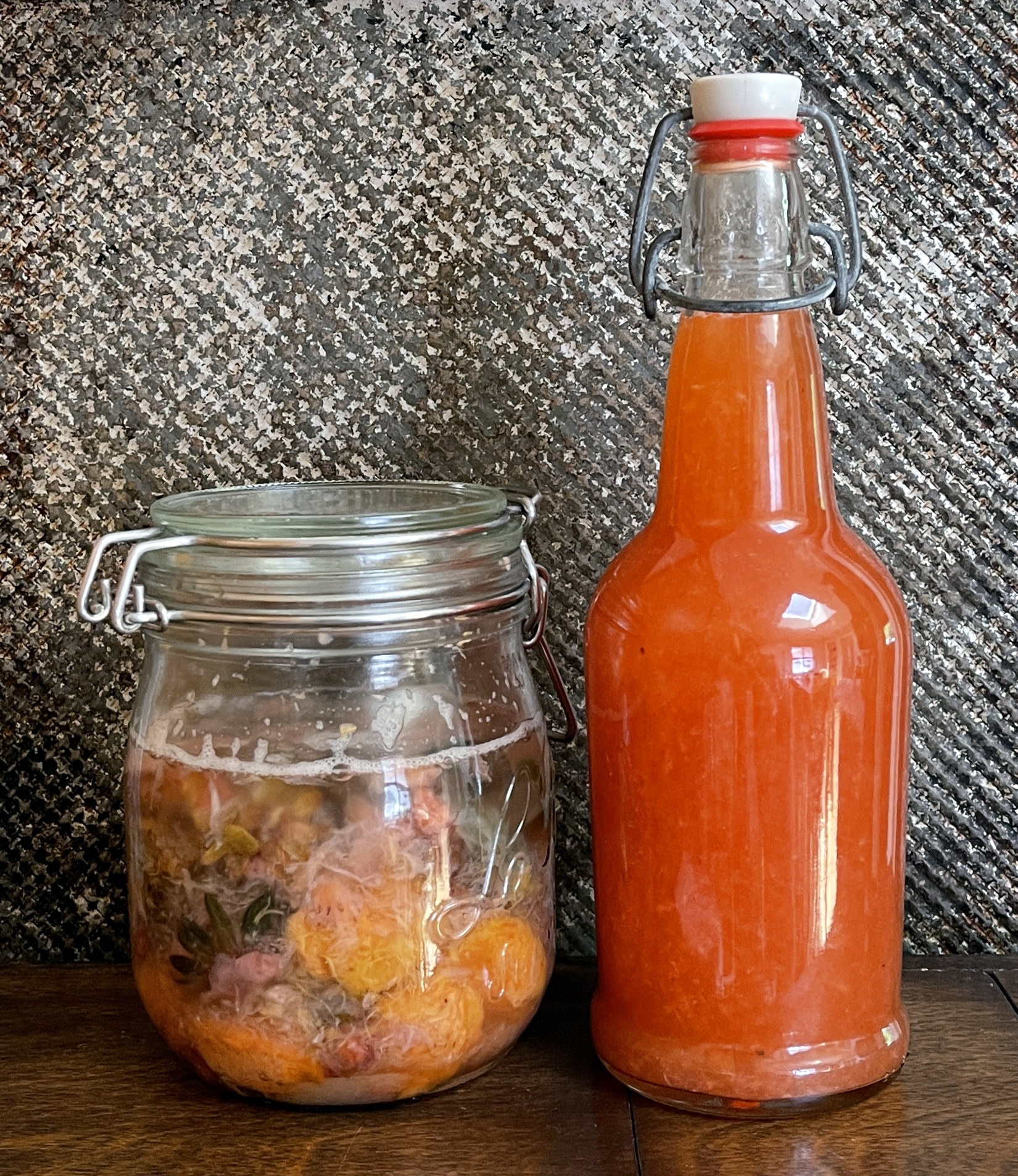 A jar of fermenting fruit scraps and a bottle of fruit kvass, ready to drink. The background is distressed grey metal. The jar and bottle are sitting on dark brown wood.