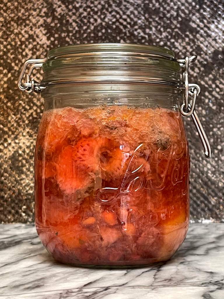 A glass jar filled with fruit scraps and water is full of bubbles during active fermentation of fruit kvass