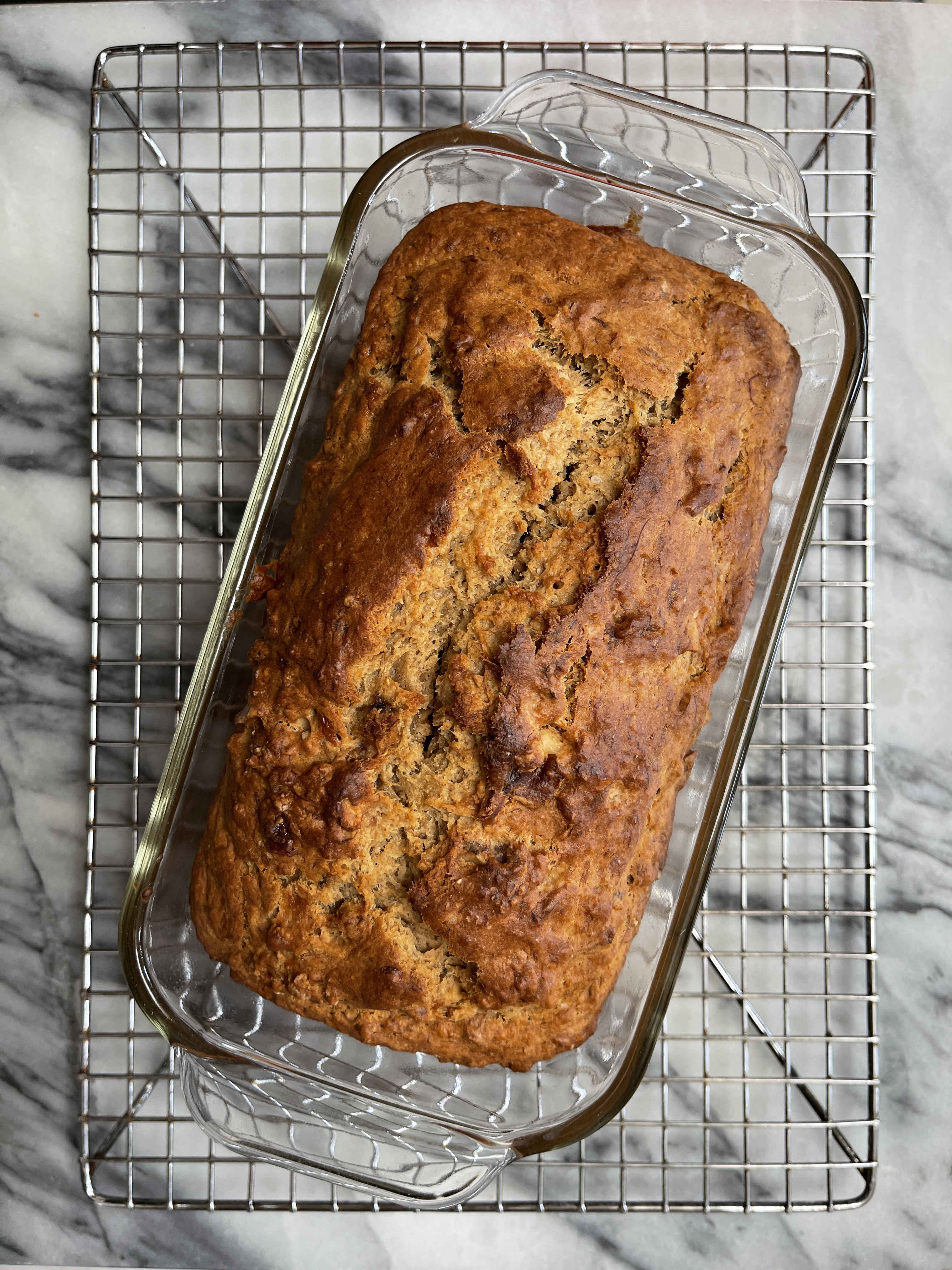 a loaf of sourdough discard banana bread cooling in a glass pan on a silver rack placed on a white and gray marble background
