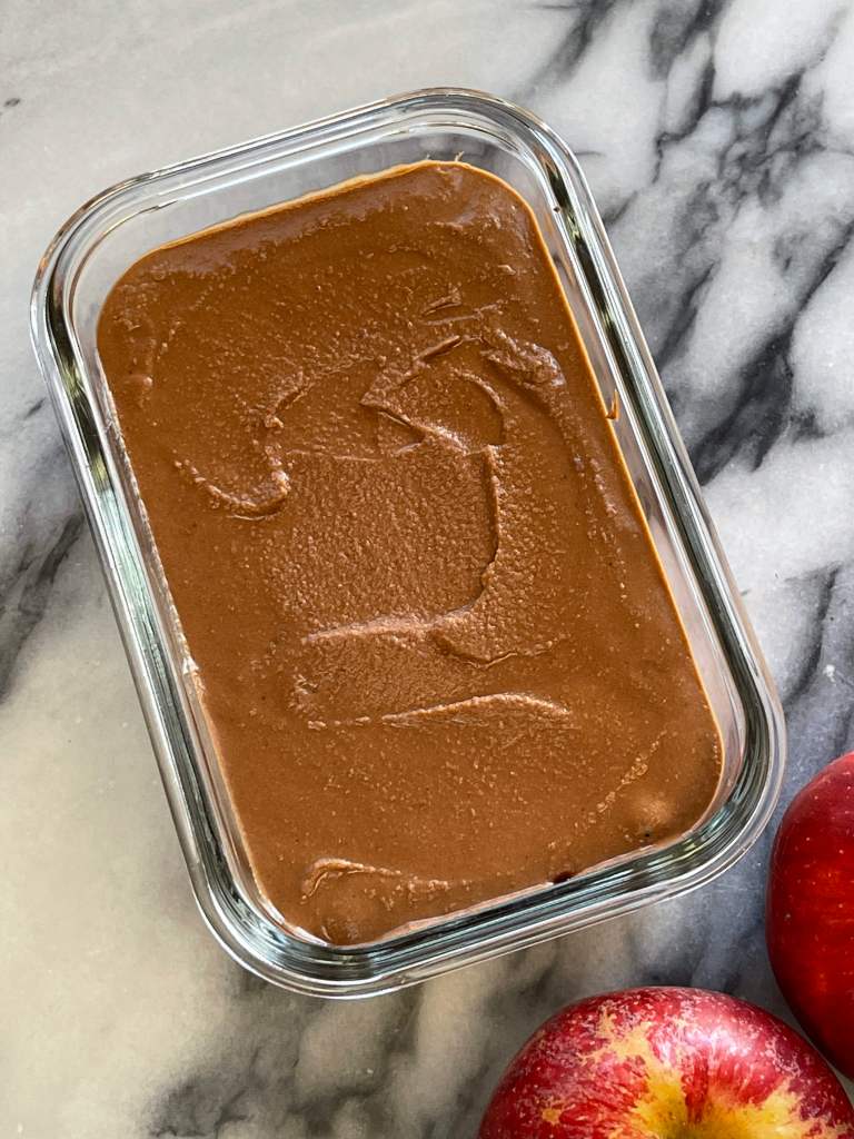 A glass dish filled with chocolate hummus sits on a grey and white marble backdrop. Two red apples sit at the bottom right of the frame.