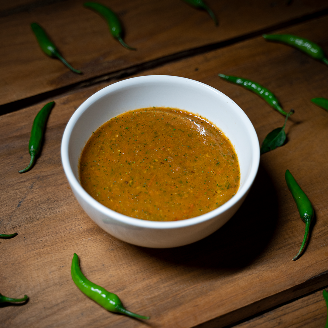 A white bowl of golveda ko achaar (tomato pickle) sits on a dark wooden table and is surrounded by green chilis