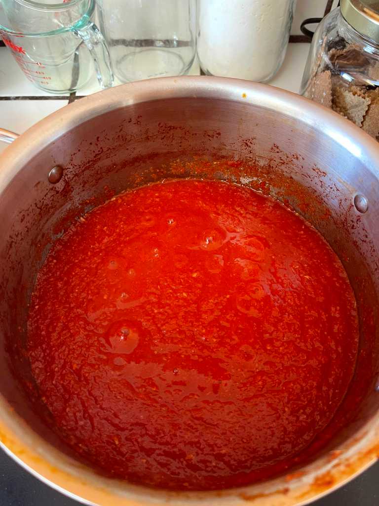 A stainless steel pot of tomato sauce cooks on an induction cooktop. In the background are several jars.
