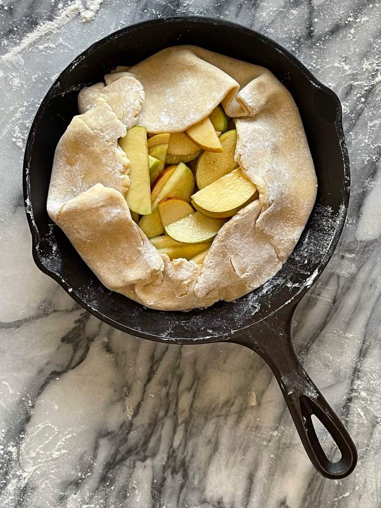 A small apple galette is ready to bake. It sits in a cast-iron pan which is placed on grey and white marble.