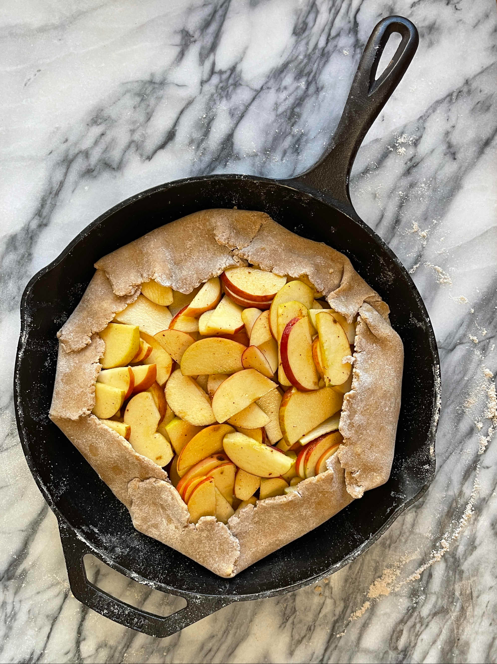 An apple galette in a cast iron pan is ready to bake. The pan sits on grey and white marble.