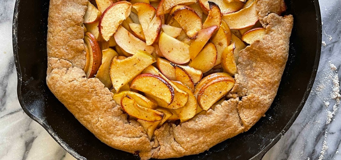 A baked apple galette in a cast iron pan sits on grey and white marble