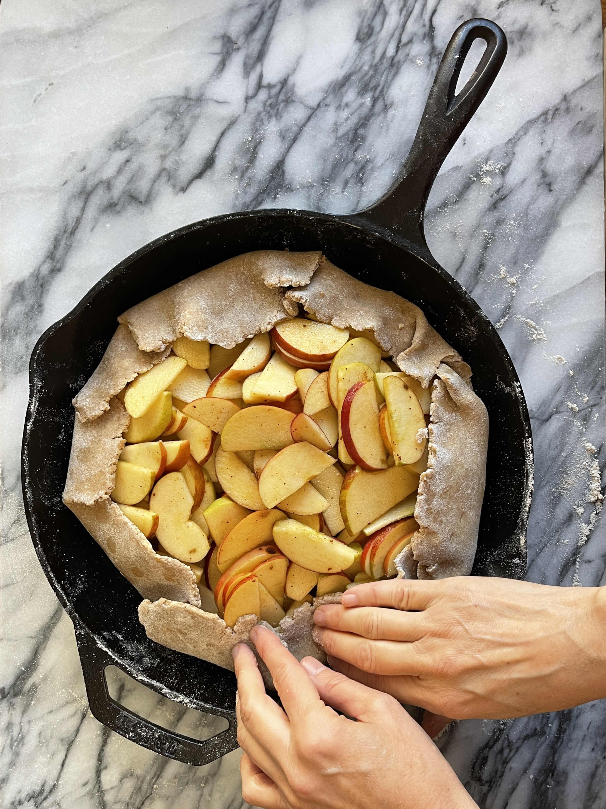 Hands fold over whole wheat dough filled with apple slices to form a galette. The galette is in a cast iron pan. The pan is sitting on grey and white marble.