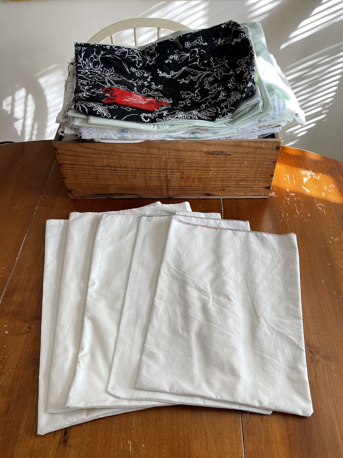 A stack of reusable cloth produce bags sits in a wooden box on a wooden table. Five white cloth produce bags are arranged on the table in front of the box.