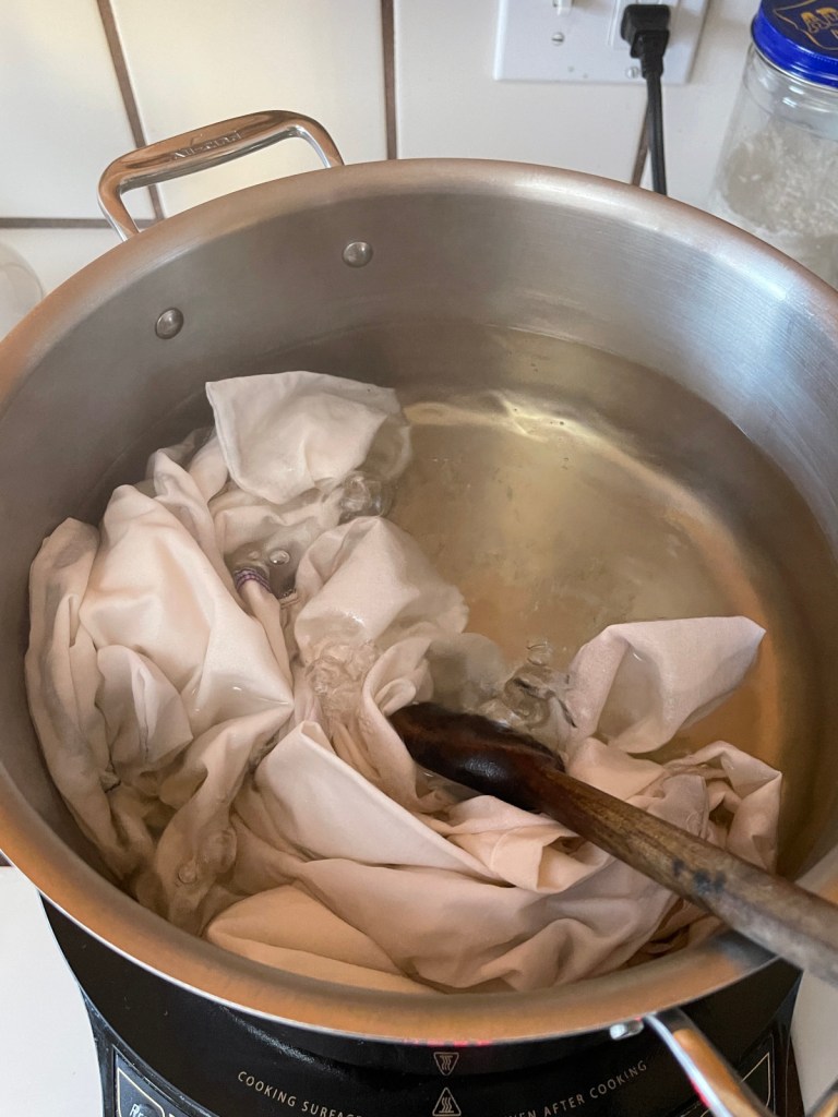 White reusable cloth produce bags simmer in a pot of water. A wooden spoon stirs them around.