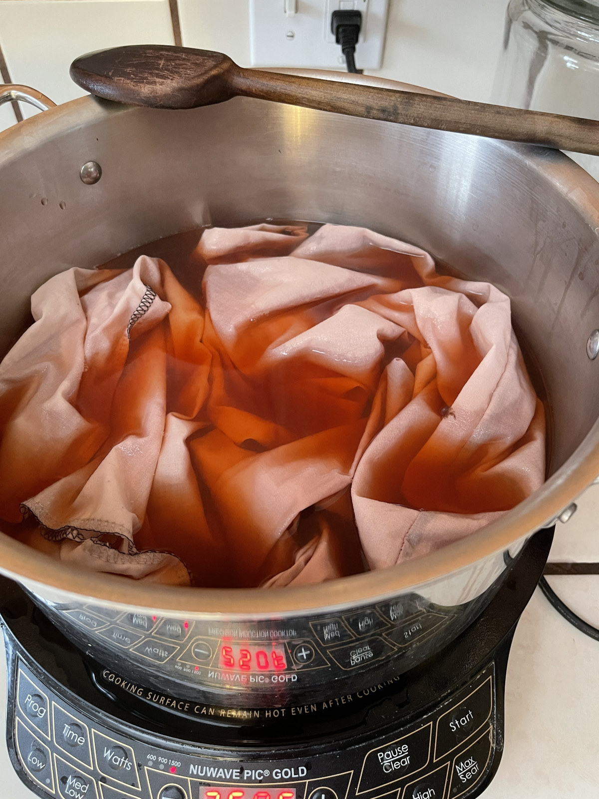 Reusable cloth produce bags sit in a large silver pot filled with avocado pit dye. The pot sits on and induction cooktop.