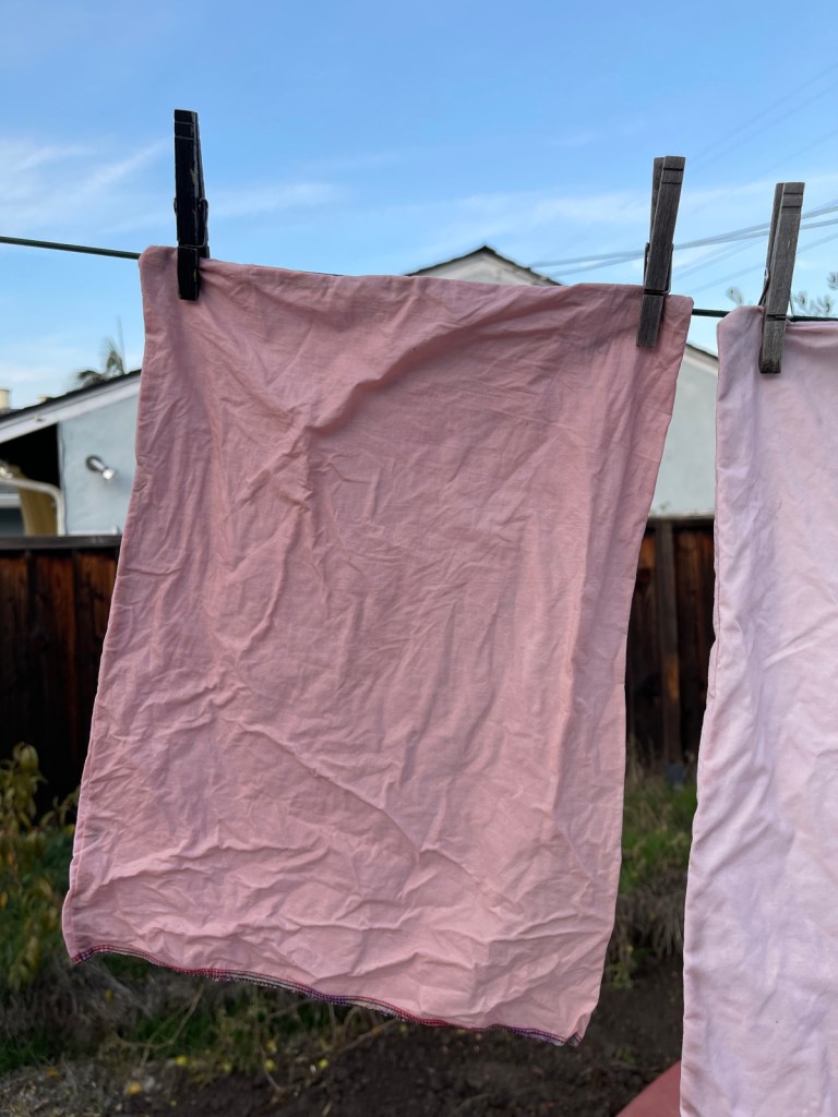 A reusable cloth produce bag dyed pink with avocado pit dye dries on a clothes line outside. Wooden clothes pins hold it in place.