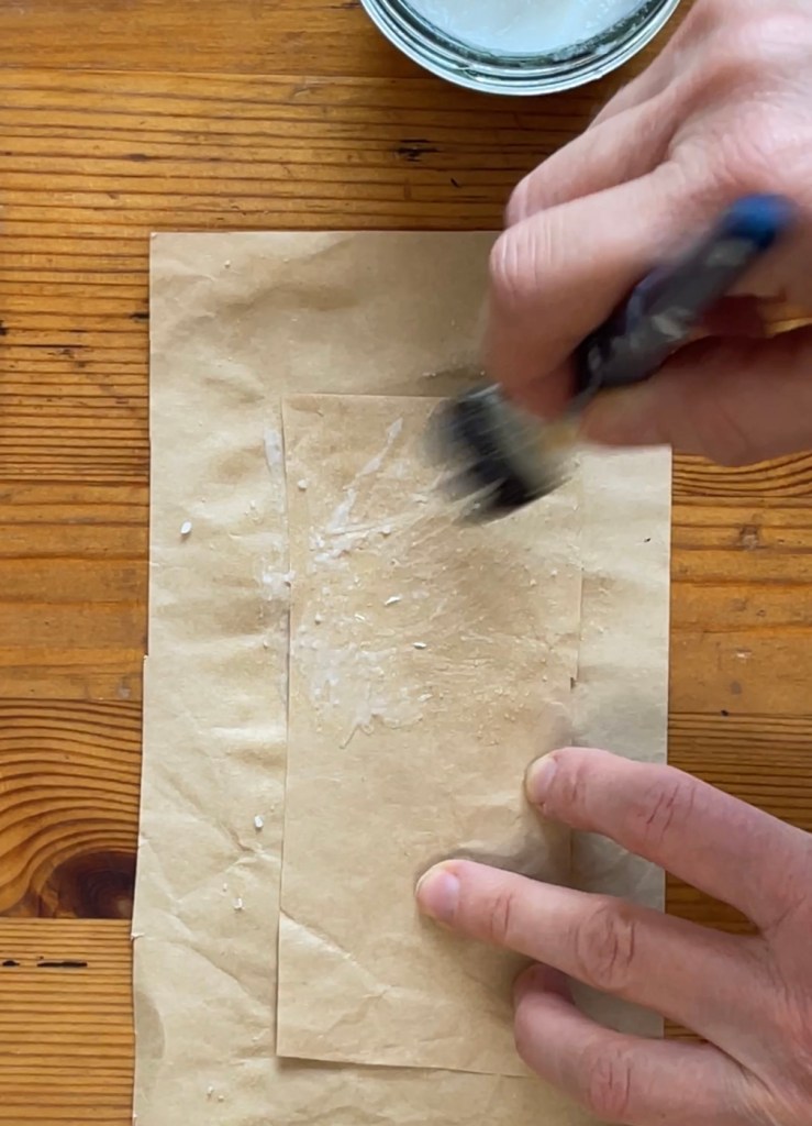 A hand holding a paint brush brushes rice glue onto a strip of brown paper tape
