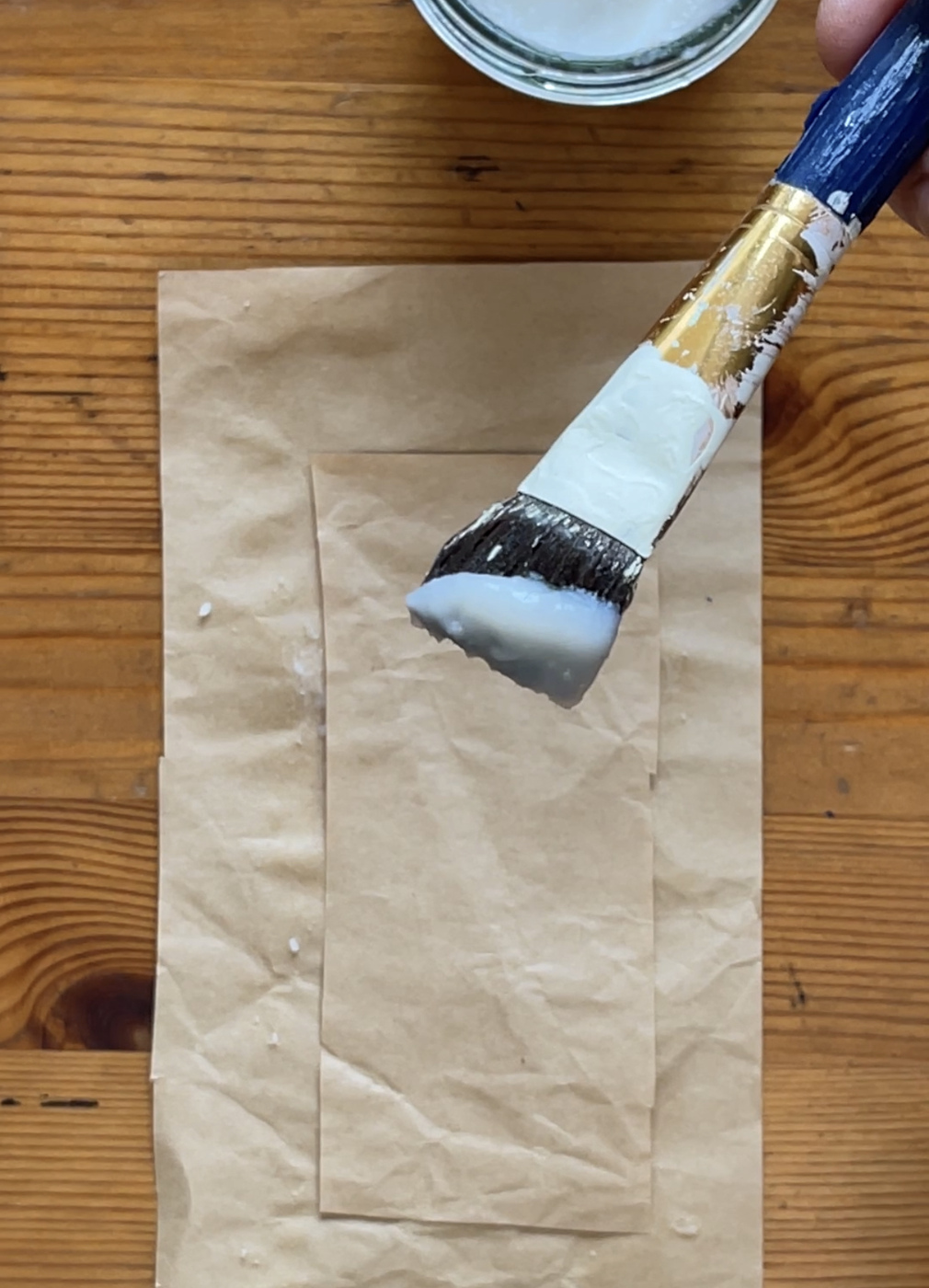 A paint brush dipped in rice glue is shown above a strip of brown paper sitting on a larger piece of brown paper.