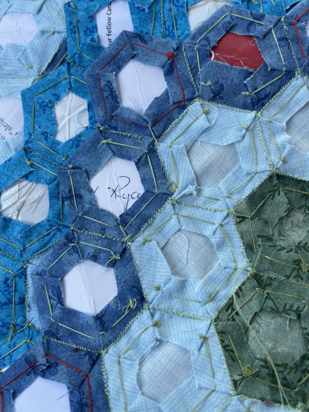 The backside of a quilt in progress shows hexagons sewn around paper. The hexagons are in various shades of blue and a bit of green.