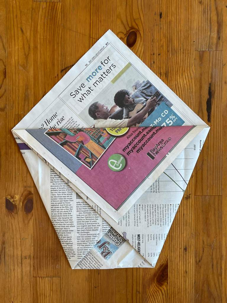 On a wooden background, a piece of newspaper is being folded into a compost bin liner.