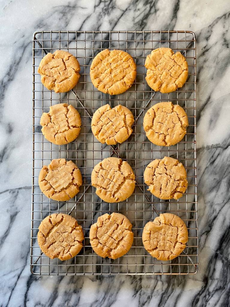 A dozen peanut butter cookies made with a sourdough discard flax egg cools in a cooling rack. The rack sits on a white and grey marble background.