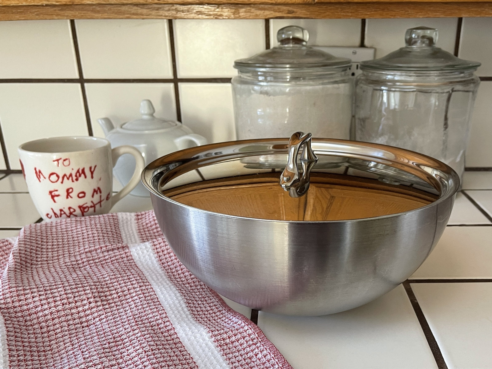 A large metal bowl of peanut butter cookie dough made with a sourdough discard flax egg sits on a white tiled counter to ferment. The bowl is covered with a metal pot lid. In the background are a red checked tea towel, a large mug, a teapot and two large jars of flour.