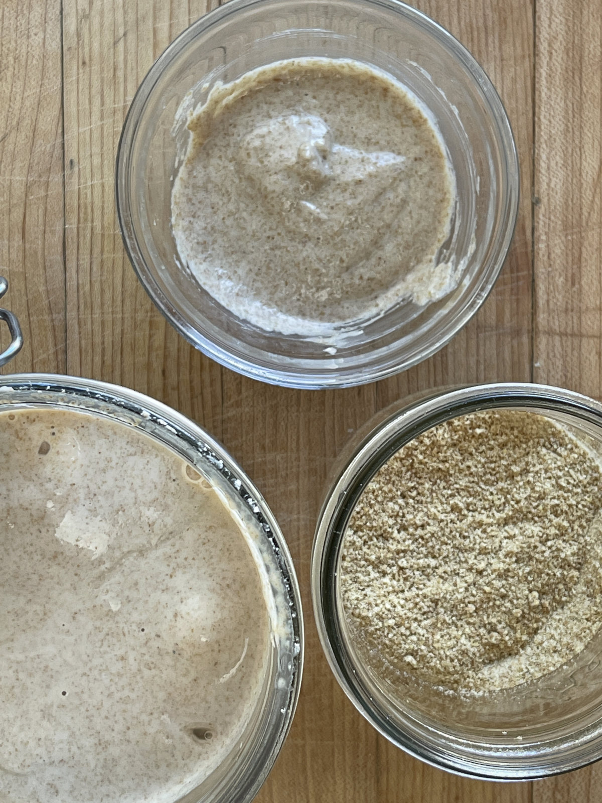 A glass bowl at the top contains a sourdough discard flax egg. The jar on the bottom left contains sourdough discard. The jar on the bottom right contains ground flaxseed meal. All three sit on a pale wooden background.