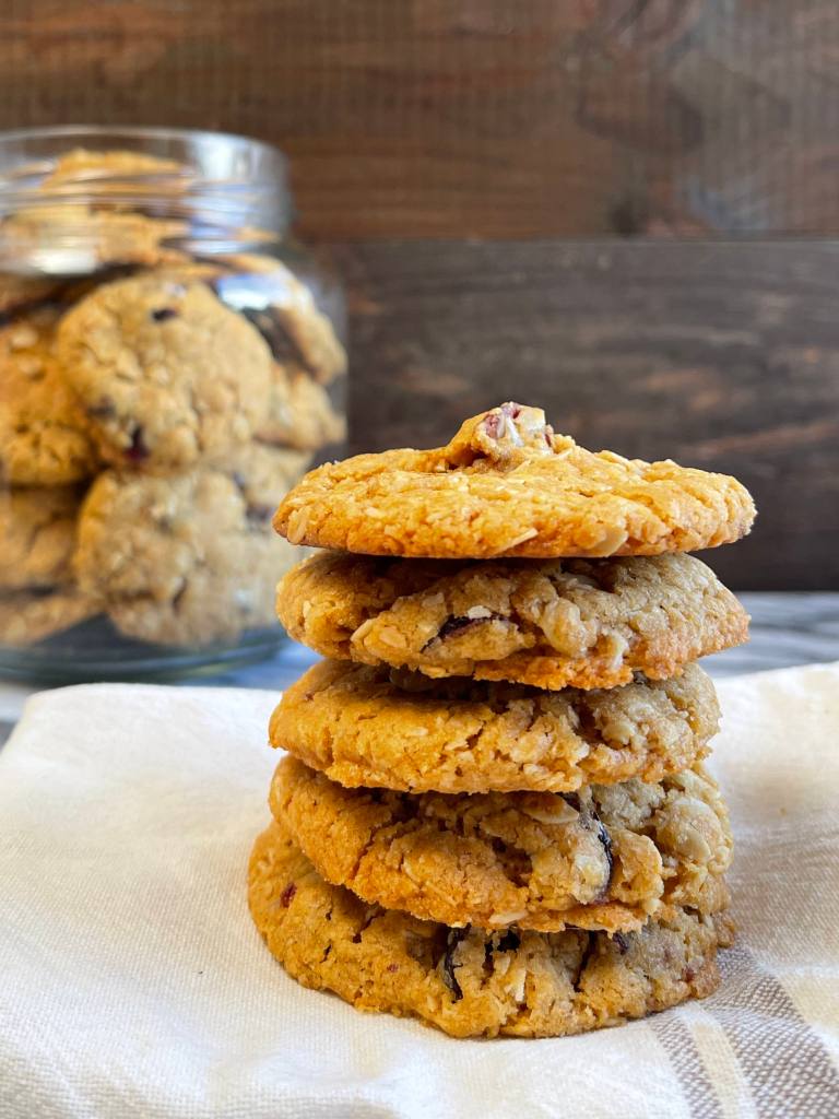A stack of 5 ranger cookies with sourdough discard sit in the foreground. In the background is a glass jar filled with the cookies