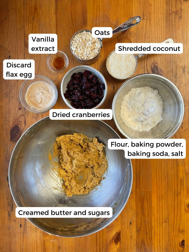 Several bowls of ingredients for ranger cookies with sourdough discard sit on a wooden table. The ingredients are labeled.