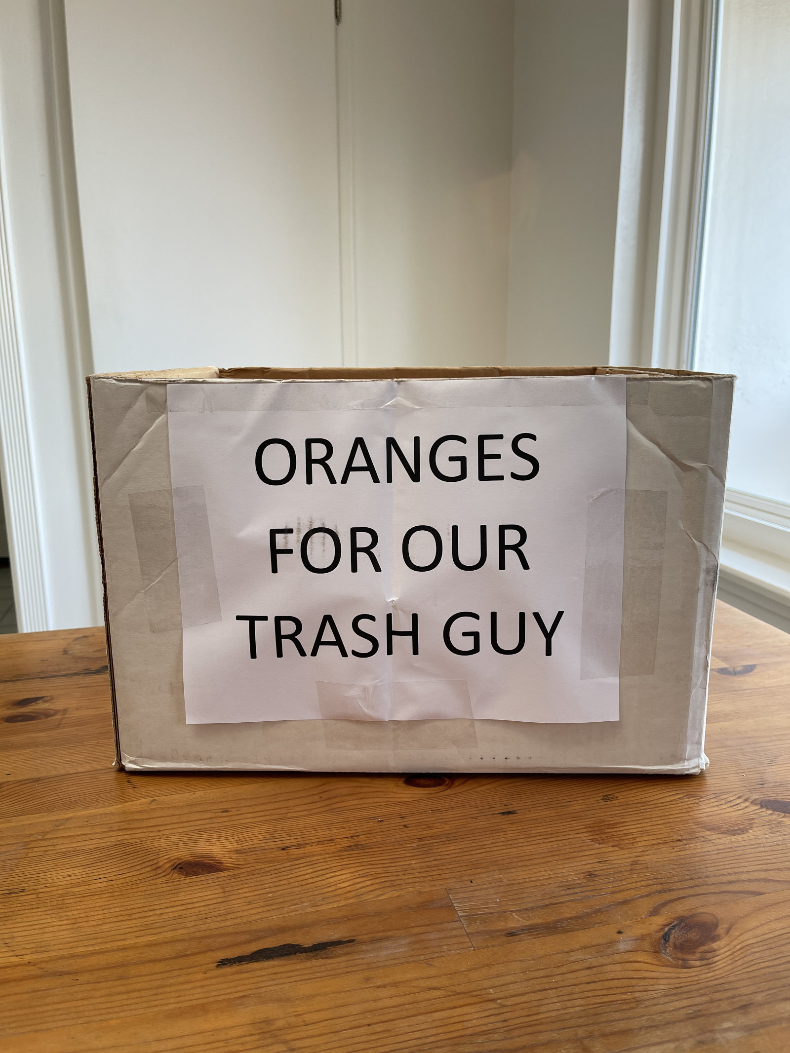 A cardboard box has a sign on the front that says "oranges for our trash guy." The box sits on a wooden table.