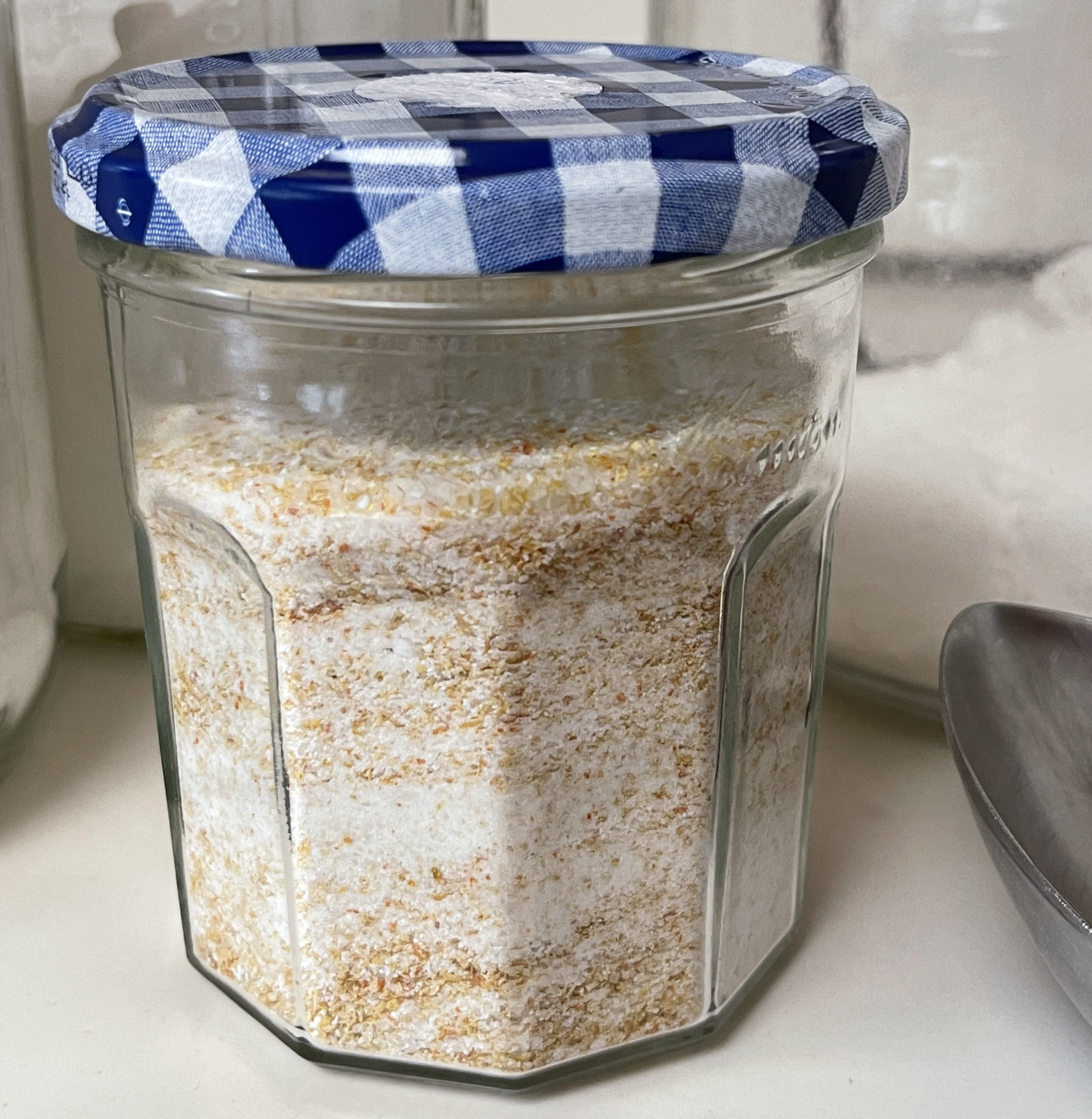 A jam jar with a blue lid sits on a while counter. The jam jar is filled with homemade scouring powder containing bits of dried citrus zest.