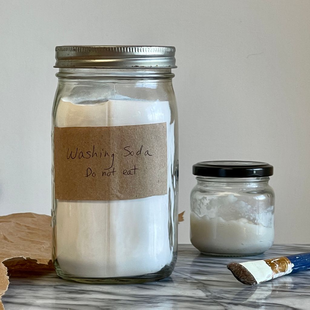 A jar of washing soda with a brown paper label sits to the left of a smaller jar half filled with wheat paste. A paint brush lies in front of the small jar.