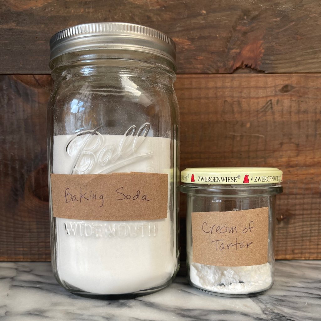 A large jar of baking soda and a small jar of cream of tartar sit on marble in front of a wooden background