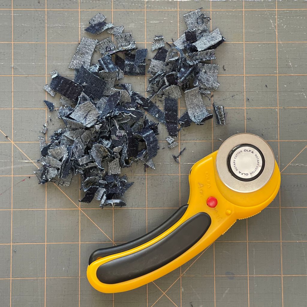 A pile of shredded dark denim sits on a gray cutting mat above a yellow rotary cutter