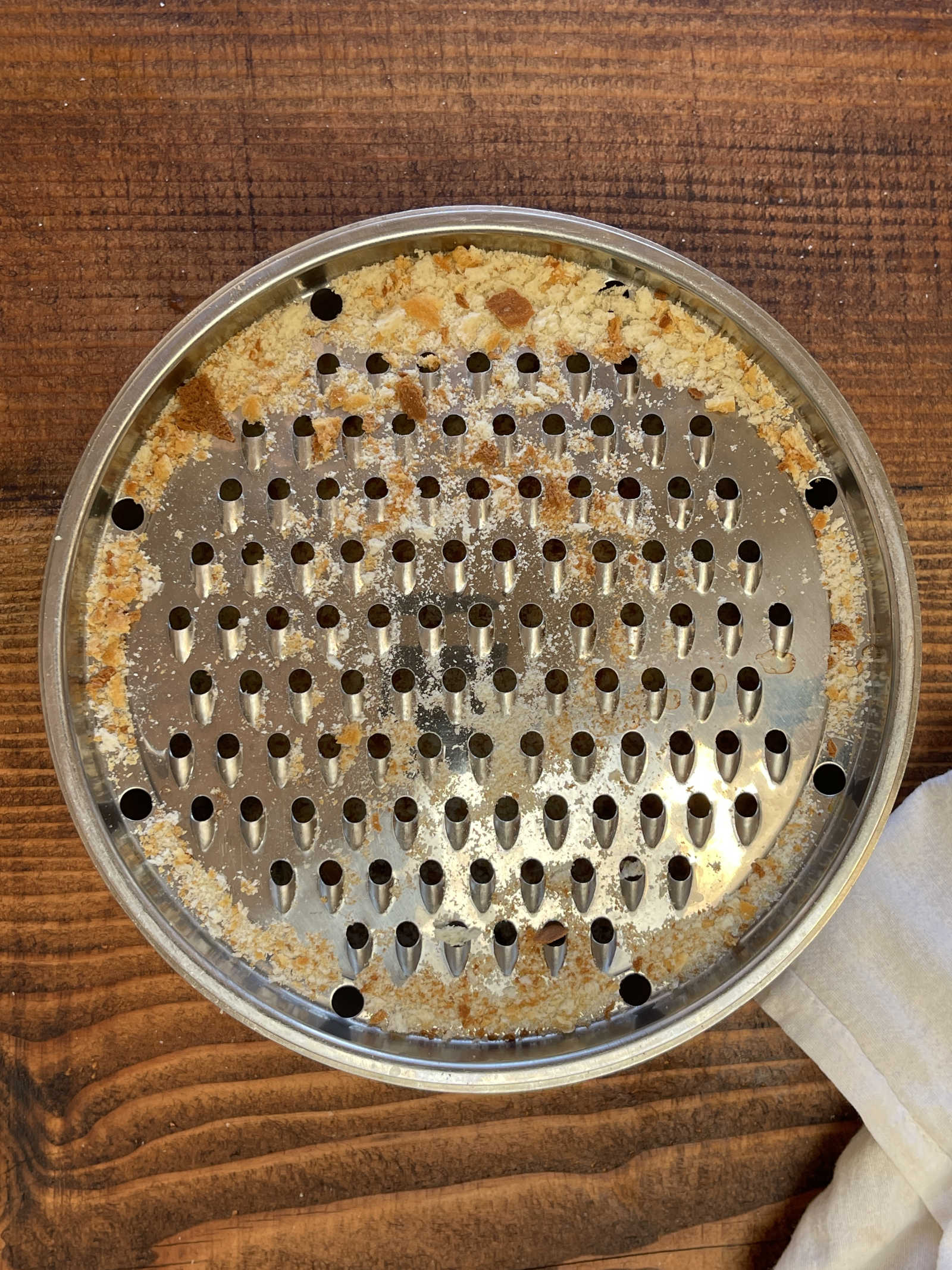 Breadcrumbs scattered on the disk of a metal cheese grater. The grater sits on dark wood.