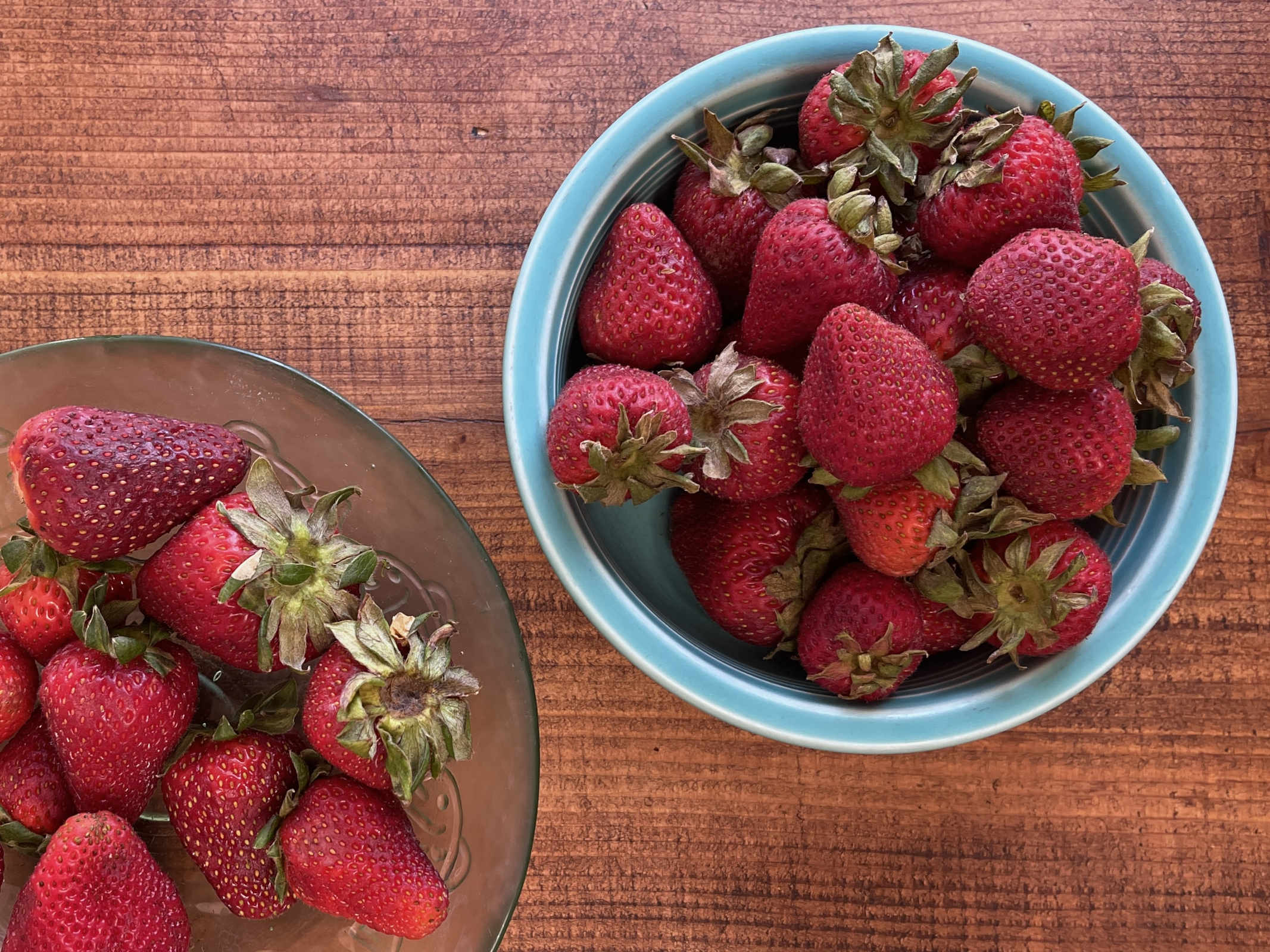 Two bowls of strawberries sit on a wooden table. They are past their peak but still edible.