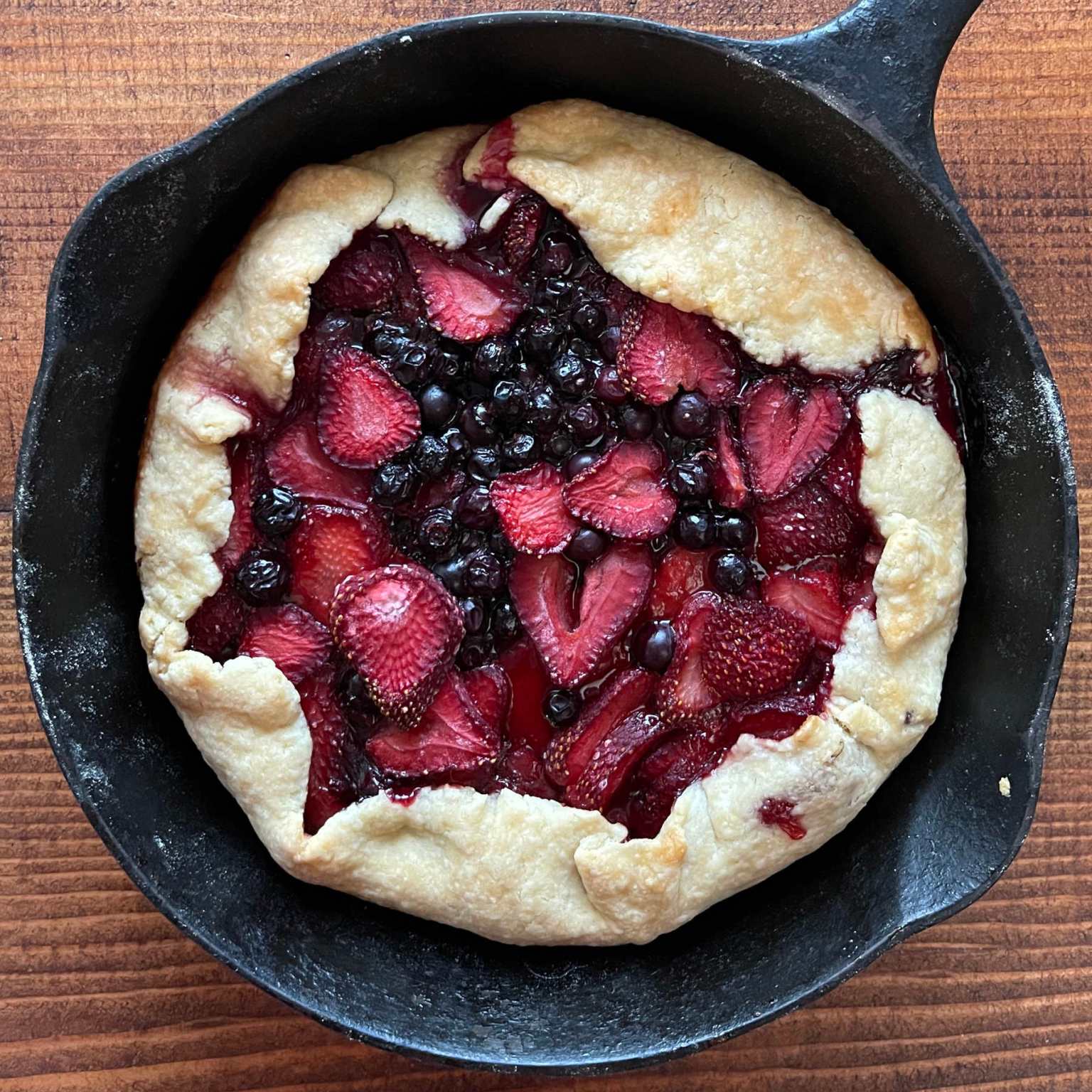 A strawberry blueberry galette baked in a cast iron pan sits on a wooden background