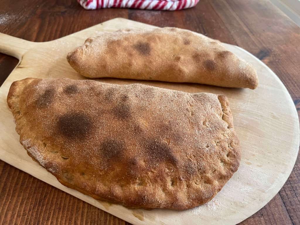 Two baked sourdough discard calzones cool on a light wood pizza peel. The pizza peel sits on a dark wooden table.