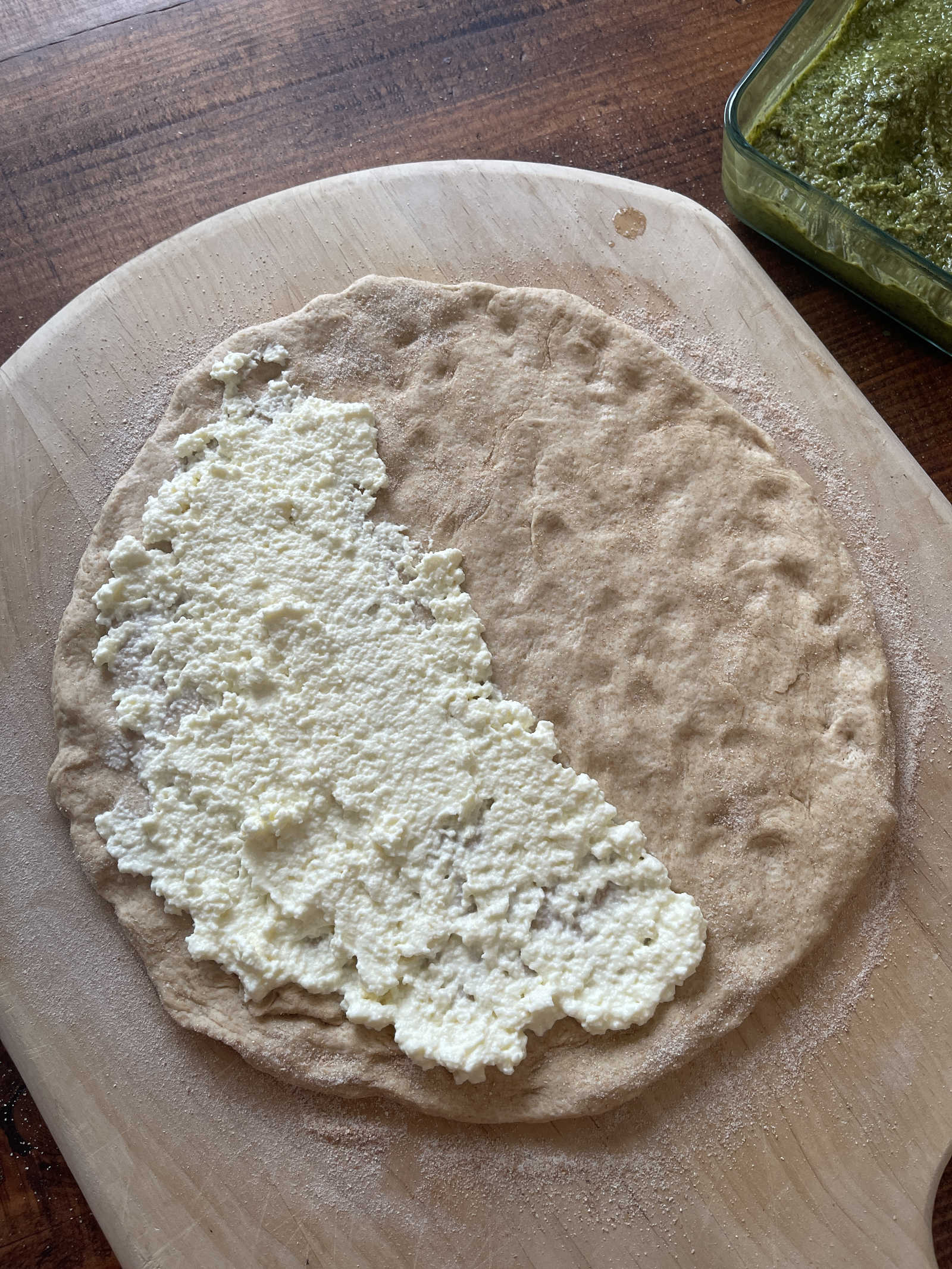 Ricotta cheese spread across half a round of sourdough discard calzone dough. The dough sits on a light colored wooden pizza peel.