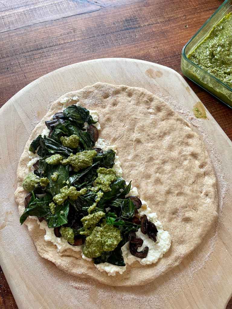 A round of pizza dough sits on a light colored pizza peel. Half of it is covered with ricotta, sautéed mushrooms, sautéed collard greens and pesto