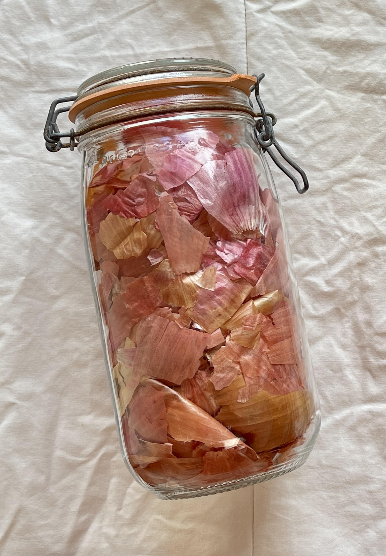 A bale-top jar filled with onion skins sits on wrinkled white fabric