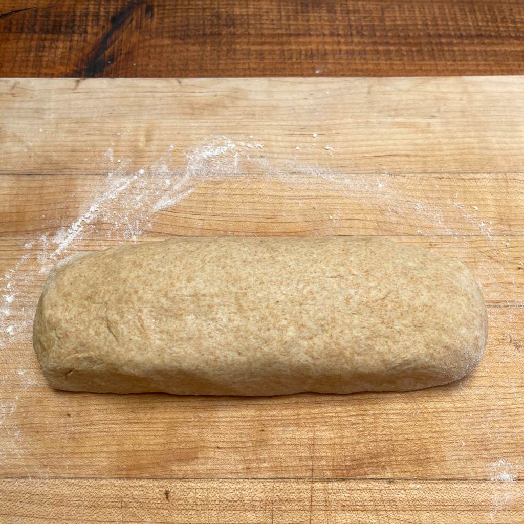 Wheat tortilla dough shaped into a log before cutting sits on a wooden board